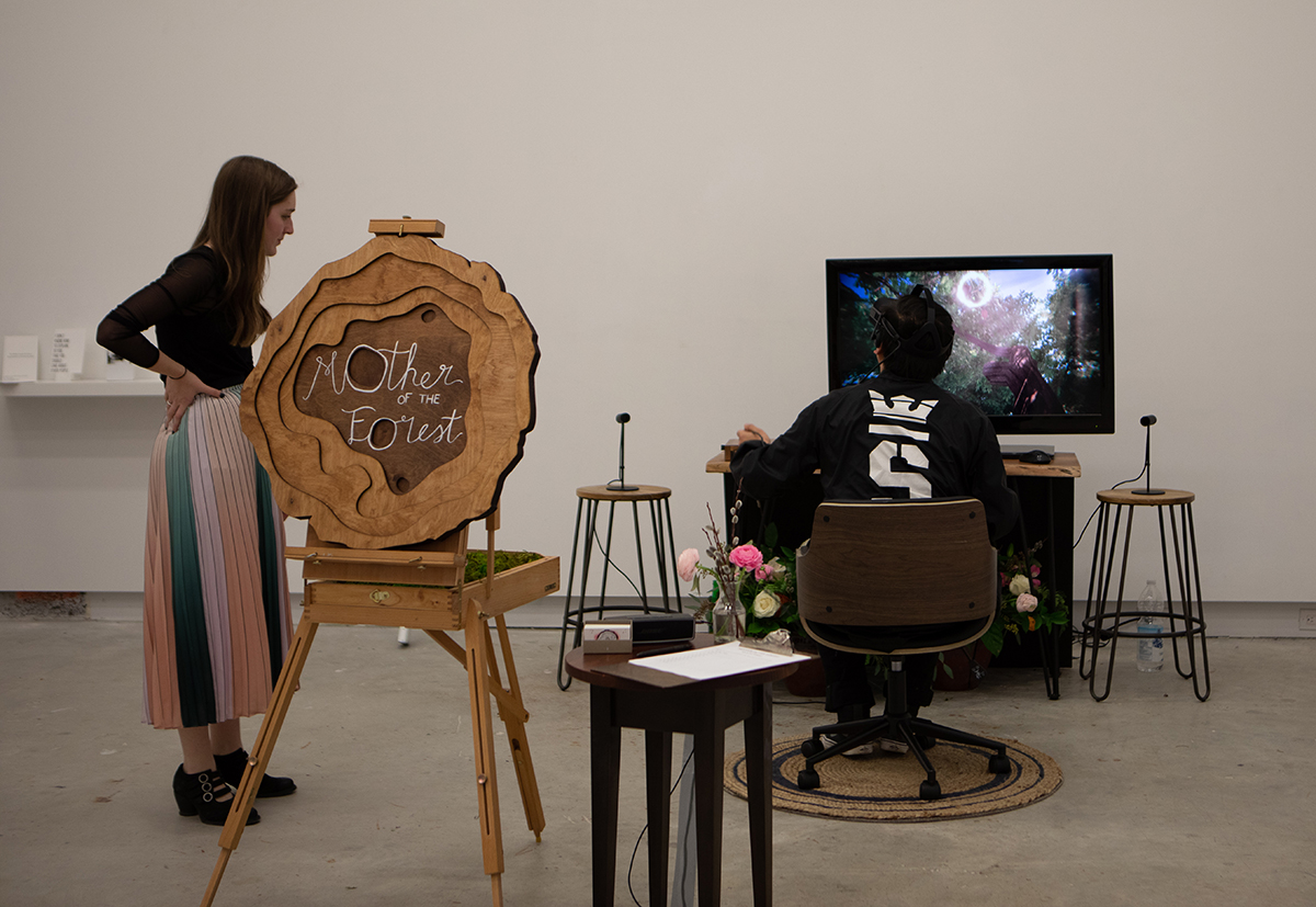 A man sitting in front of a TV screen wearing a VR headset. Next to the man is a woman and a wooden sign that reads 'Mother of the Forest'.
