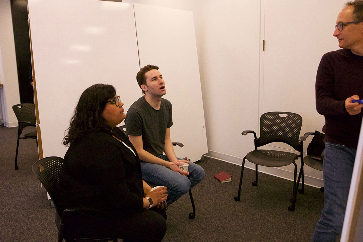 Two people sitting on chairs while one person is standing next to a white board.