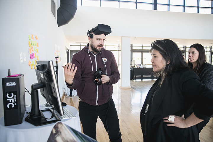 A man with a VR headset around his head is talking to two women as they stand next to him and look at a computer screen.