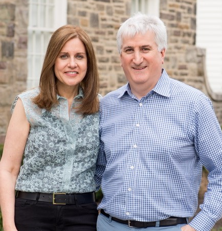 Two people standing side-by-side in front of a brick house