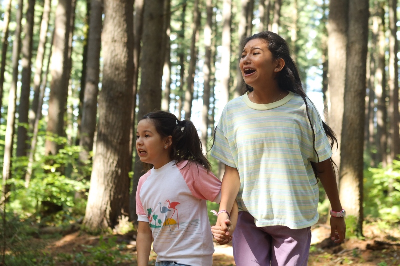 Two sisters with distressed looks on their faces hold hands while standing in a forest