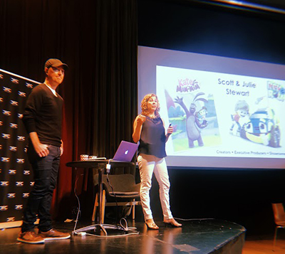 A man and woman stand onstage, with a screen projecting animated character images to their right.