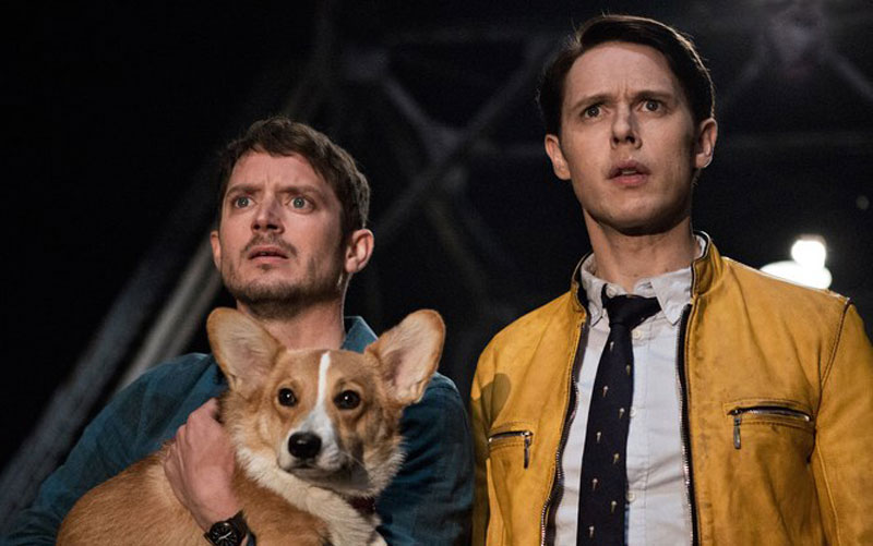 Two men who are standing side by side each other, the man on the left is holding a corgi dog, and both of them have shocked expressions on their faces.