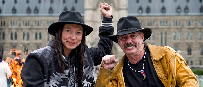 Two Indigenous folks stand together while holding up their fists in solidarity 