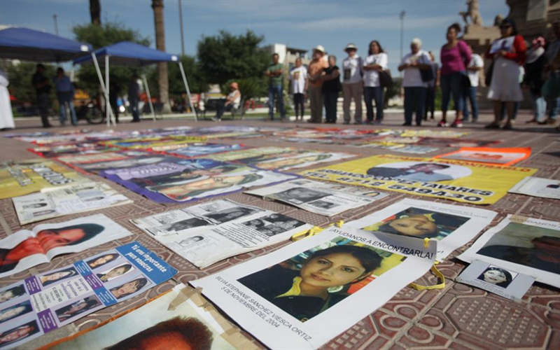 A still shot of the ground covered in pictures of people, with people surrounding them and looking at them.