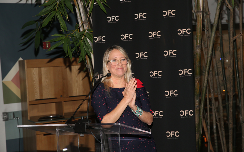 A blonde female standing on a podium at an event introducing the next speakers.
