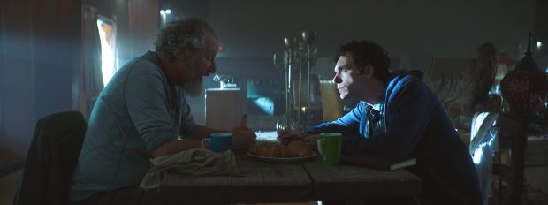Two men sit across a table from one another in a dark room