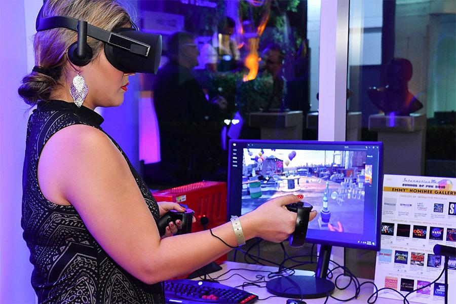 A woman wearing a VR headset and holding a controller while standing in front of a computer monitor.