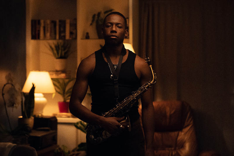A man wearing a black tank top holds a saxophone, standing in the middle of a dimly lit room.