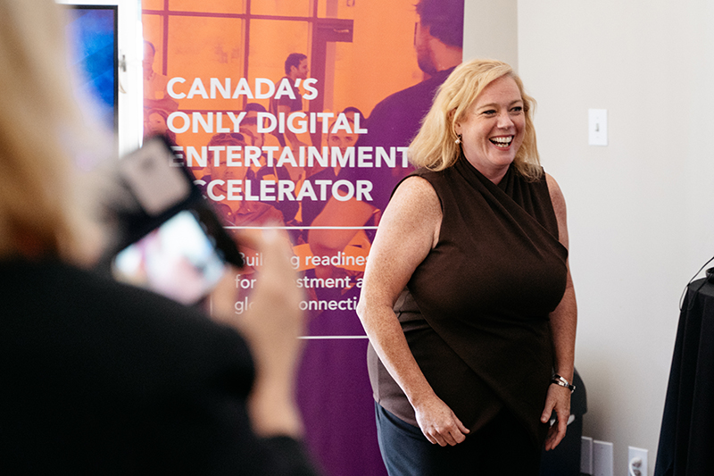 A woman smiling, standing in front of a banner that has the words "Canada's Only Digital Entertainment Accelerator."