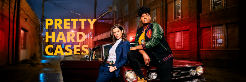 Two female detectives, one leaning against the hood of a car and one sitting on the hood of a car, posing for a promotional picture for a tv series