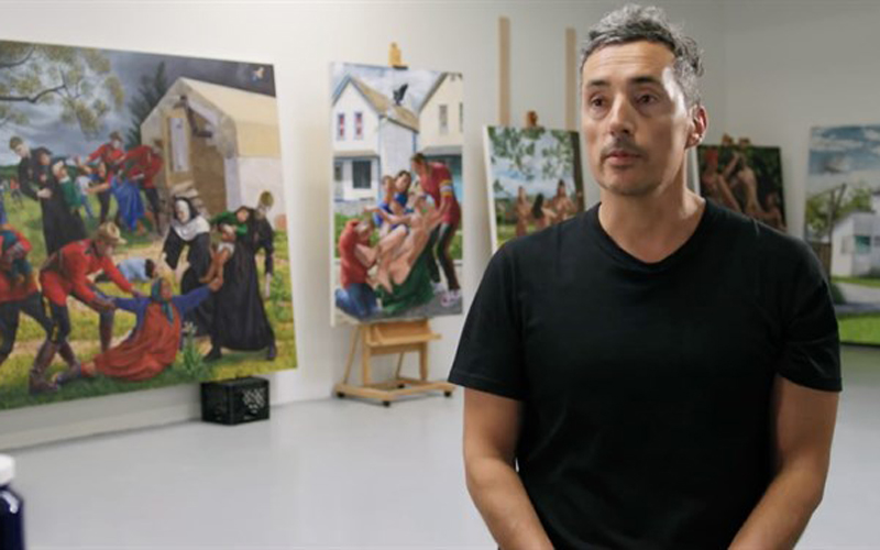 A still image of a man standing in front of a gallery talking to the camera