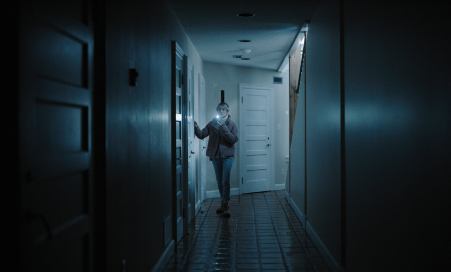A woman wearing a coat, jeans and boots walks down a dark hallway with lots of doors, using a flashlight to guide her