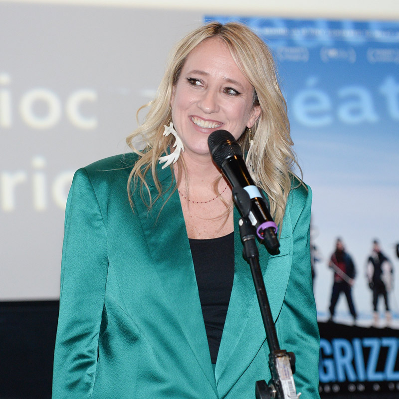 A blonde female in a green silk suit talking into the microphone on stage.