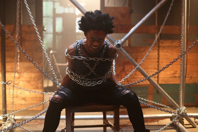 A woman with an angry look on her face sits on a chair, tied down by several chains