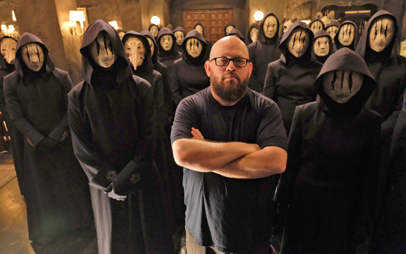 A still photo from a TV show of a man standing in glasses, surrounded by a crowd of people in masks. 