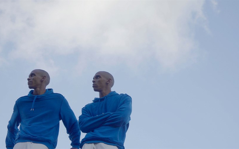 A image of two males that are twins, standing by each other wearing the same blue sweater, with the clear sky in the background.