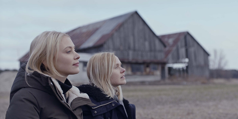 Side profile of two sisters standing outside wearing coats in front of a barn looking at something outside of the frame