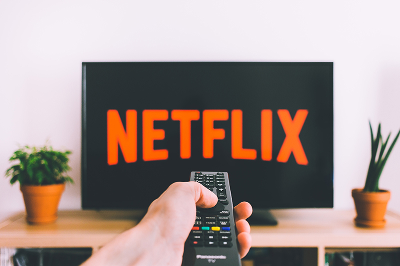 A television with the 'Netflix' logo on it. In front of the TV is a hand holding a remote.