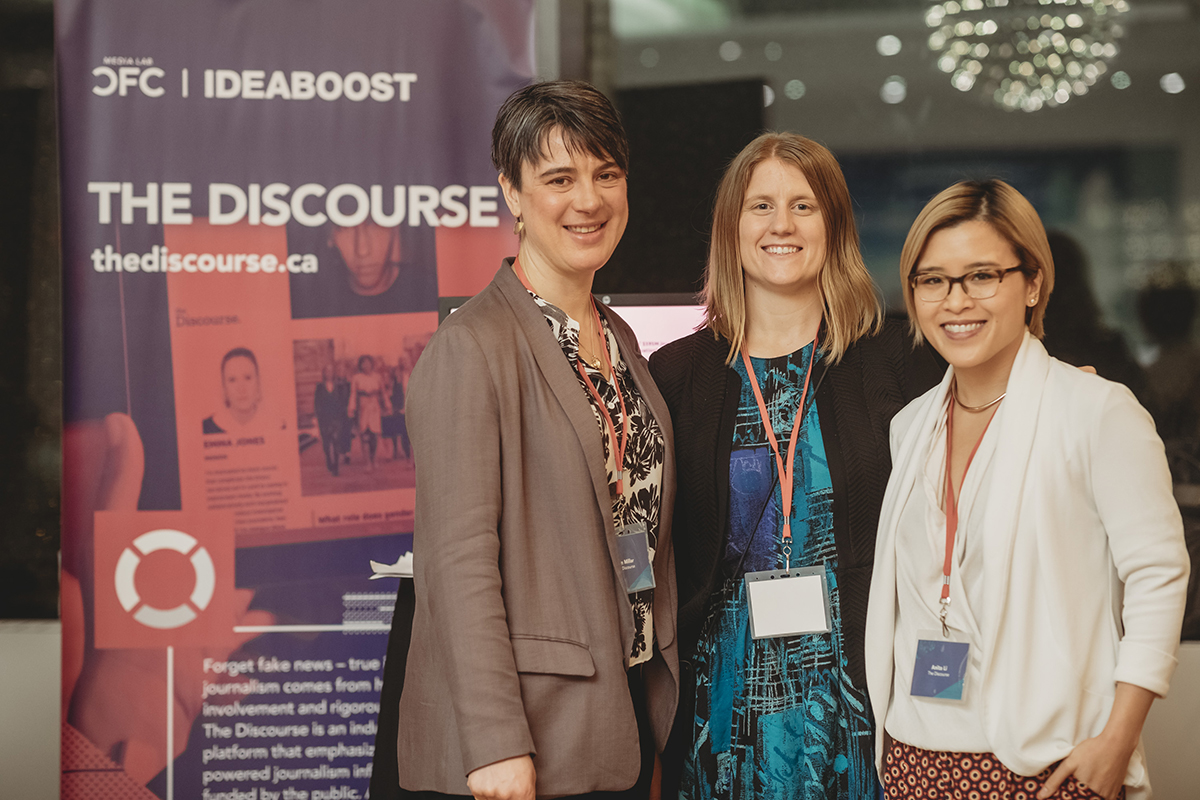 Three women standing next to a pink pull up banner for 'The Discourse'.
