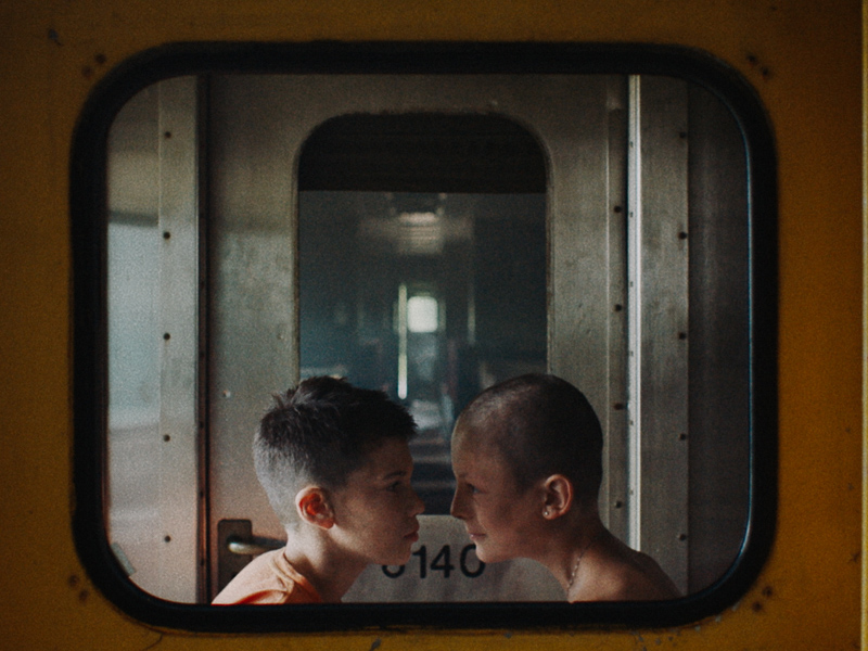 Two boys face each other against a dirty industrial set of doors and windows.