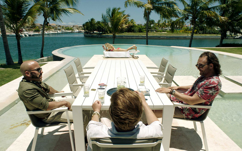 A still image of three men sitting at a poolside table talking to each other over some drinks, and a lady laying on her back on the tanning chair by the pool in a distance. 