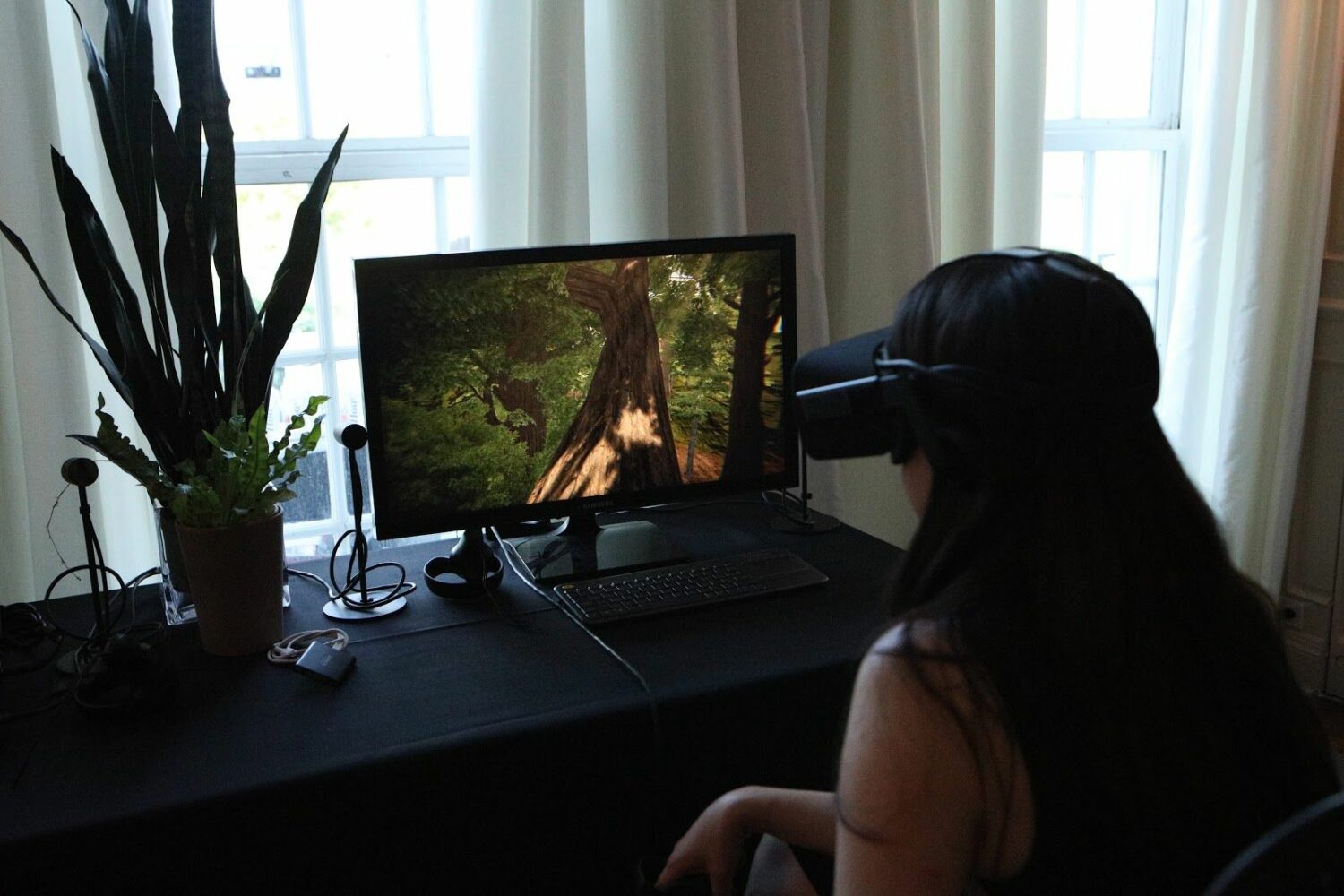 A person sitting at a desk wearing a VR headset. On the screen in front her is an image of a forest. 