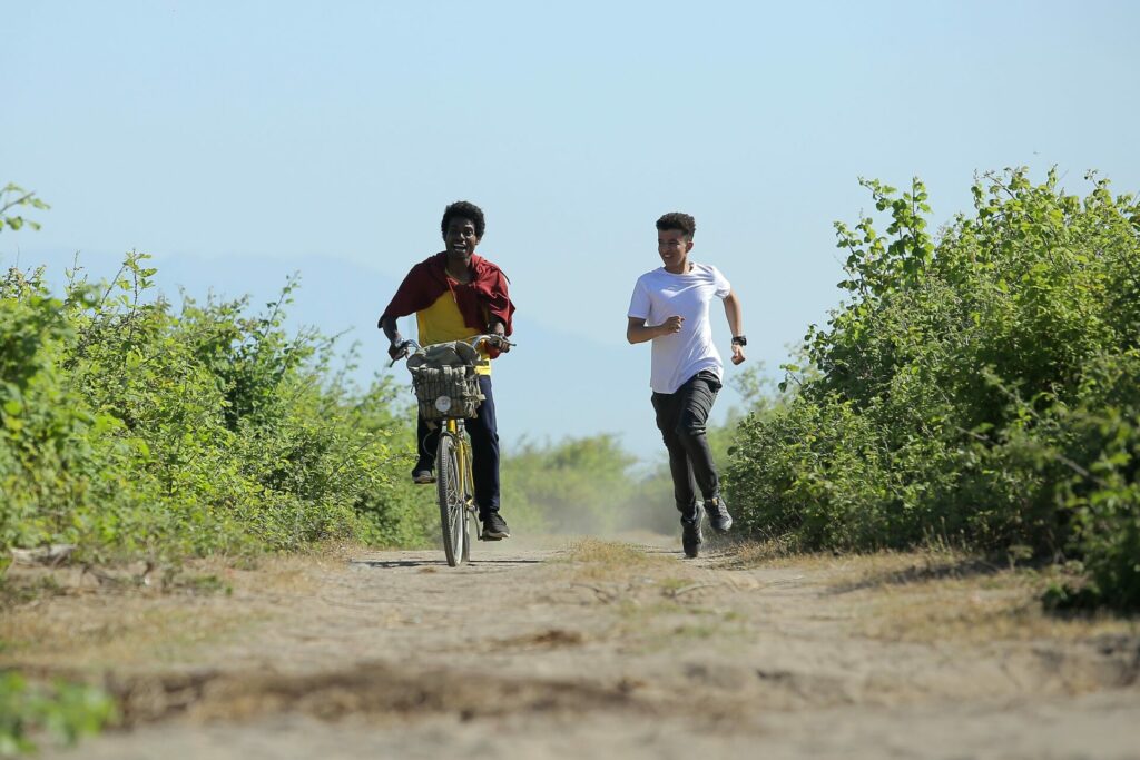 A young adult riding a bike outdoors with another young adult running beside him.