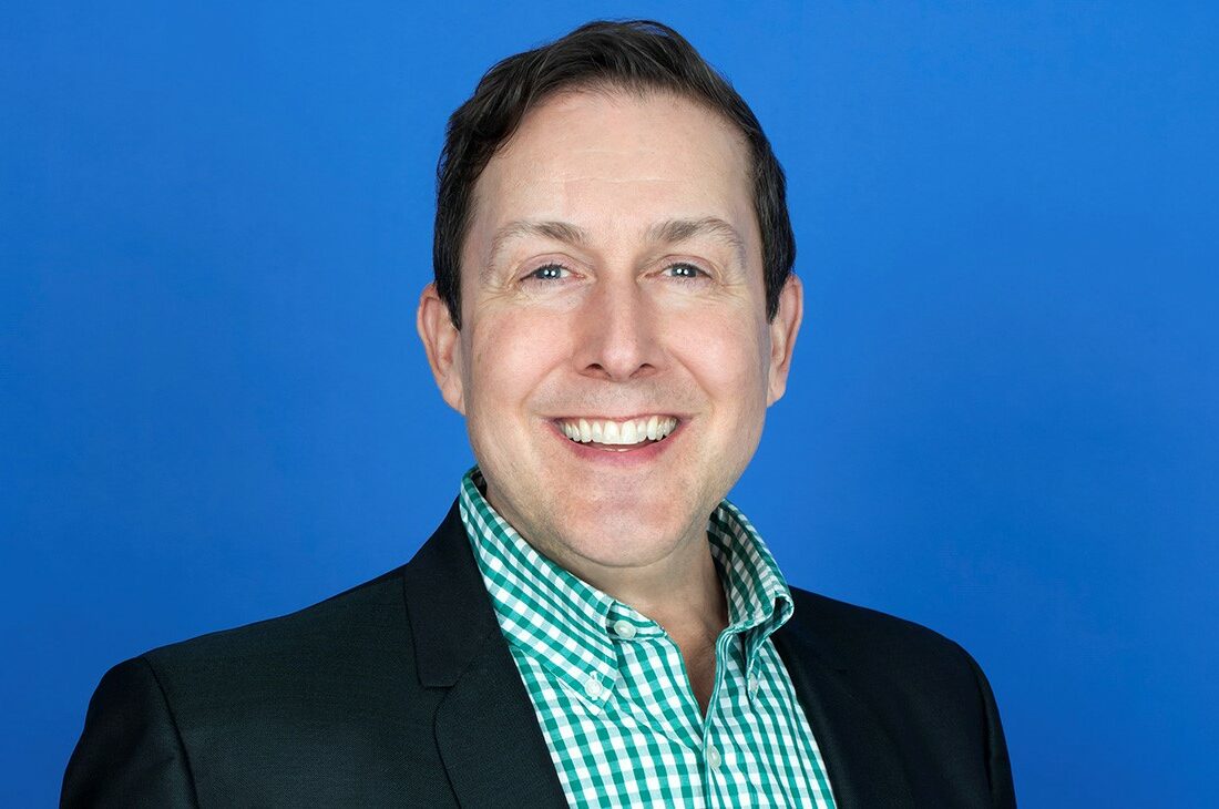 A headshot of a man wearing a green checkered shirt and a black blazer in front of a blue background.