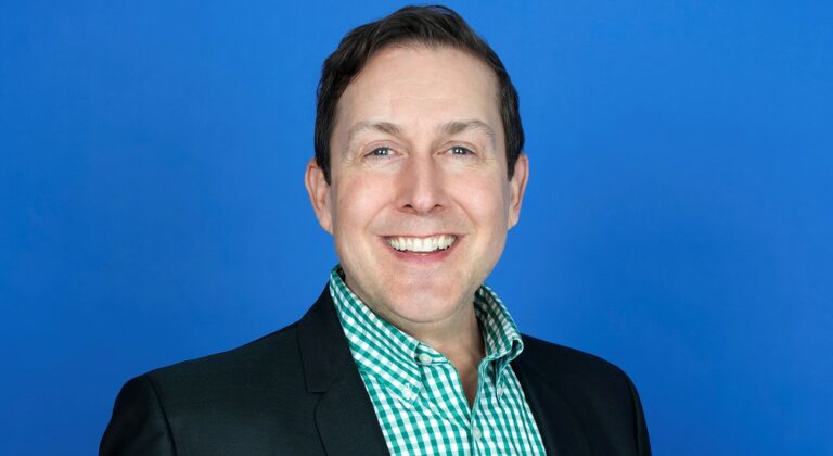 A headshot of a man wearing a green checkered shirt and a black blazer in front of a blue background.
