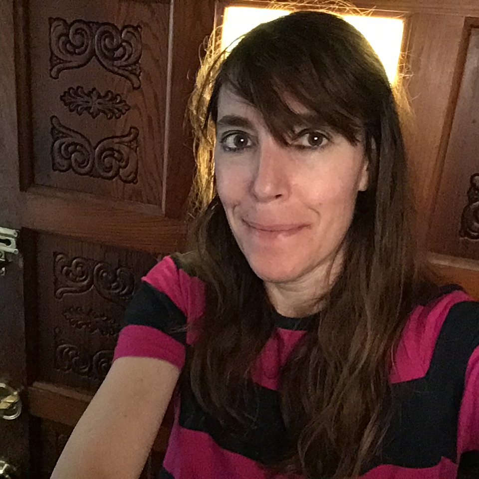 A selfie of a woman with brown hair and bangs wearing a pink and black striped t-shirt.